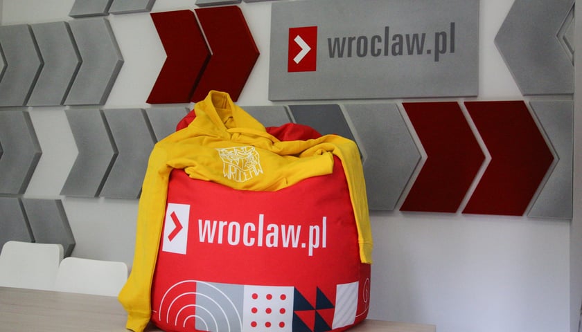 Czerwona pufa Wroclaw.pl i leżąca na niej żółta bluza z herbem Wrocławia. Wszystko na tle napisu na ścianie Wroclaw.pl