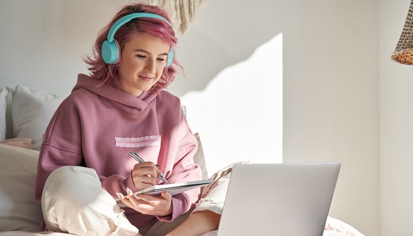 Młoda osoba w bluzie siedzi na łóżku przed laptopem, na uszach ma słuchawki, a w dłoniach notatnik i długopis, lekko się uśmiecha (zdjęcie ilustracyjne) 