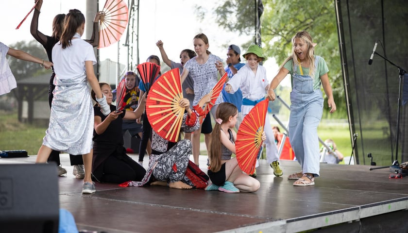 Scena na świeżym powietrzu, na scenie grupa nastolatków wykonuje układ z dużymi czerwonymi wachlarzami. W środku trzy osoby kucają, pozostałe dzieci stoją  