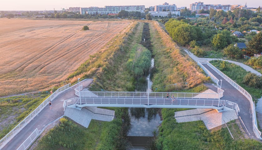 Kładka nad rzeką w otoczeniu zieleni, na niej dwójka rowerzystów, obok spacerująca osoba