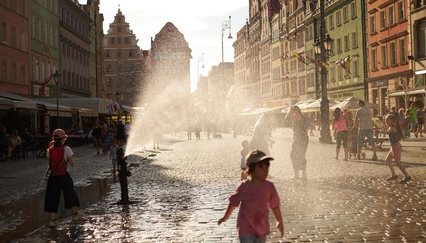 Rynek, brukowana ulica, z hydrantu leci woda, tworząc pióropusz kropel. Wokół biegają dzieci 