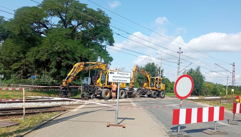 Remont przejazdu przez tory na ulicy Głównej/Maślickiej. Widać dwie koparki oraz białą tablicę z napisem "Przejście wyznaczonym korytarzem"