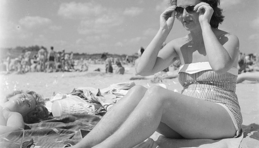 Wakacje nad Bałtykiem w PRL-u. Młoda kobieta siedzi na plaży w kostiumie kąpielowym i okularach. Rok 1958