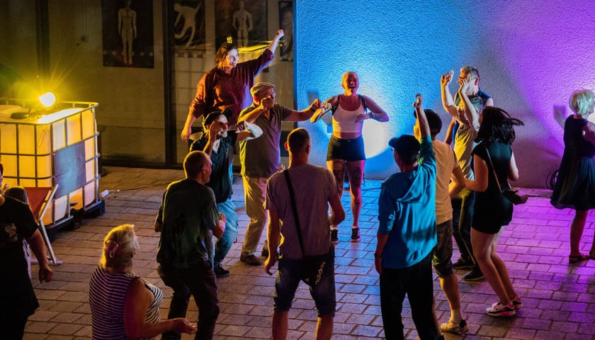 Potańcówka na Oławskim. Na zdjęciu mieszkańcy okolicy tańczący na podwórzu Centrum na Przedmieściu przy ulicy Prądzyńskiego. Zdjęcie wykonane po zmroku, imprezie towarzyszy kolorowe oświetlenie. 