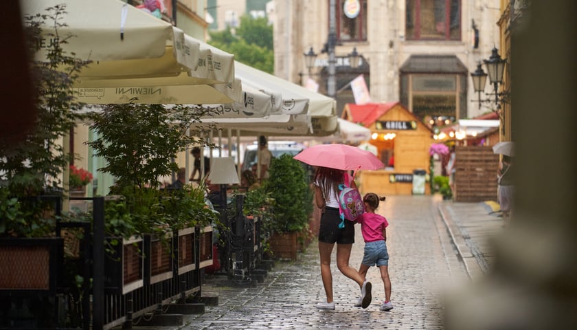 Ogródki gastronomiczne na wrocławskim Rynku, przy których idzie kobieta z dzieckiem schowane przez deszczem pod parasolką. 