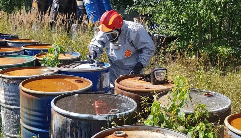 Ubrany w kombinezon ochronny człowiek stoi między dużymi beczkami i pobiera próbkę do badania  