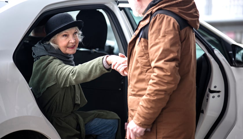 Seniorzy przy samochodzie. Mężczyzna ubrany w brązową kurtkę pomaga wysiąść z samochodu kobiecie w czarnym kapeluszu i zielonej kurtce 