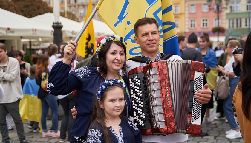 Kobieta w granatowej bluzce z haftem, mężczyzna z nałożoną akordeonem i dziewczynka w granatowej bluzce z haftem. W tle tłum   