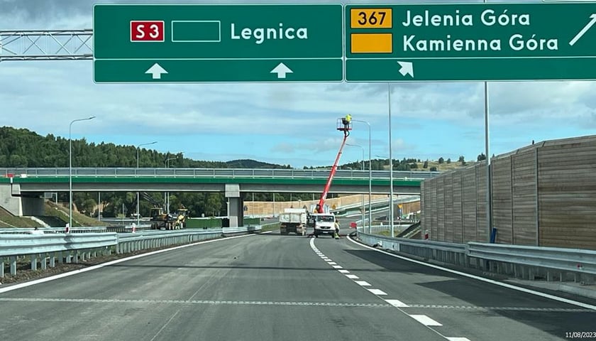 Ulica, a nad nią zielone tablice z napisami. Na jednej: S3 Legnica, na drugiej: Jelenia Góra, Kamienna Góra  