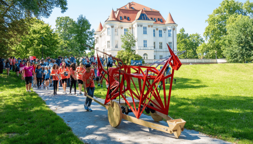 Korowód dzieci. Na pierwszym planie ażurowa rzeźba zwierzaka, którą pcha na wózku chłopiec. W tle zabytkowy duży budynek, trawnik i park
