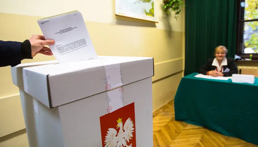 Dłoń osoby wrzucającej formularz do głosowania do urny wyborczej, w tle osoba siedząca przy stole. Zdjęcie ilustracyjne