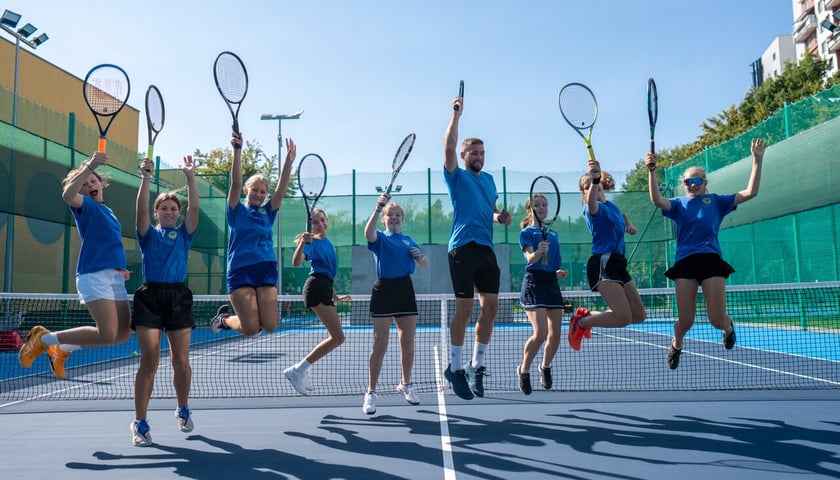 Ośmioro  dzieci i jeden dorosły, wszyscy ubrani w niebieskie koszulki i czarne spodenki,  skaczą do góry z podniesionymi nad głową rakietami tenisowymi 