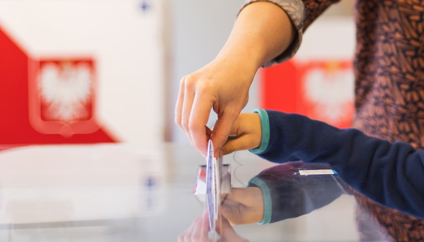 Biało-czerwona urna do głosowania. Zbliżenie na dwie dłonie, które wrzucają do niej białą kartę 