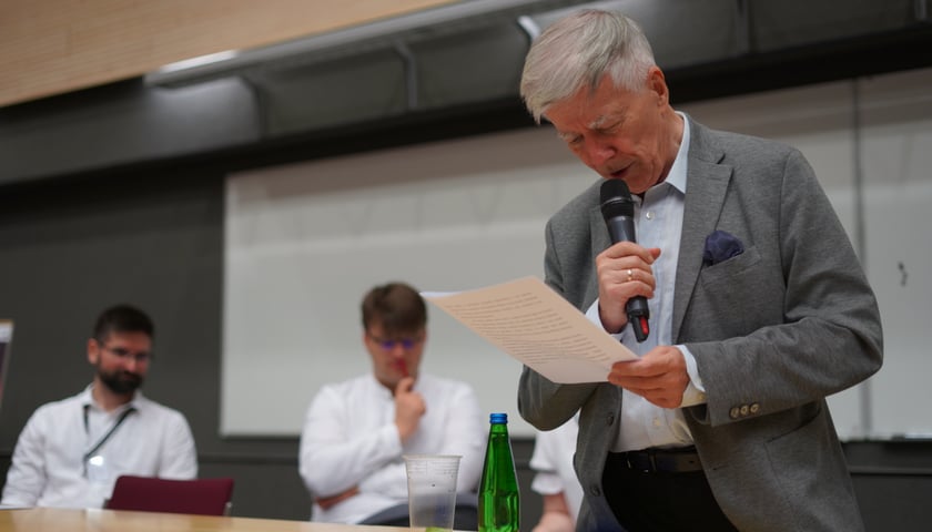 Pochylony nad kartką prof. Jan Miodek trzyma mikrofon i odczytuje dyktando na sali uniwersytetu