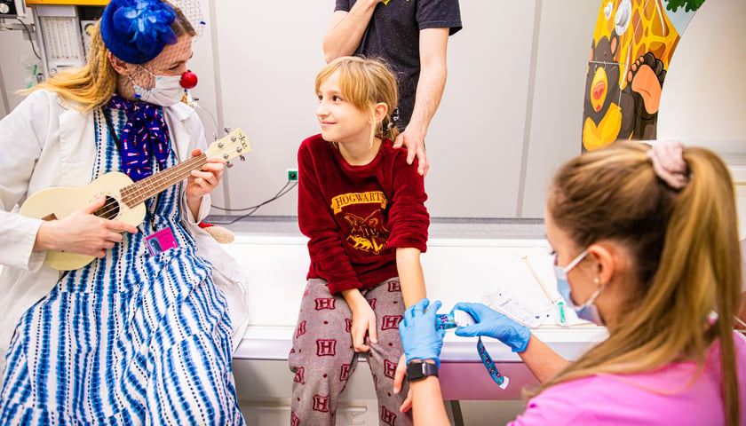 Na szpitalnym łóżku siedzi dziewczynka w bordowej piżamie, uśmiecha się lekko, patrząc na kobietę klowna, która gra na małej gitarze. Obok pielęgniarka szykuje zastrzyk    