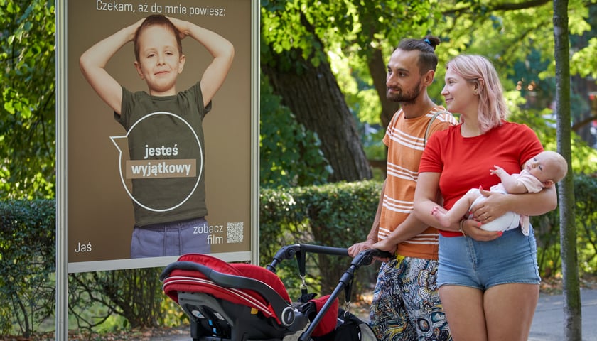 Rodzina, kobieta w czerwonej bluzce i spodenkach z niemowlakiem na rękach i mężczyzna w pomarańczowej koszuli w paski, pchający wózek, patrzą na planszę, na której widać chłopca. Napis na planszy: Czekam, aż do mnie powiesz: jesteś wyjątkowy. Jaś  