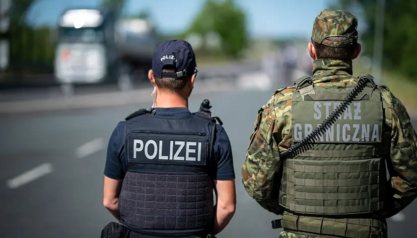 Dwie osoby stojące tyłem, ubrane w mundury z napisami "policei" i "straż graniczna" 