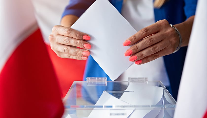 Na zdjęciu damskie dłonie trzymające złożoną kartę do głosowania nad przezroczystą urną wyborczą