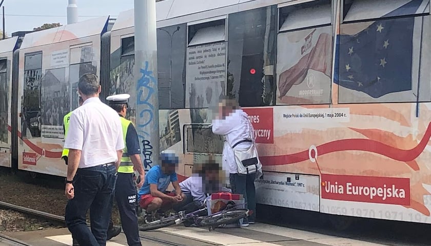 Mężczyźni i kobieta, w tle tramwaj i przewrócony rower 