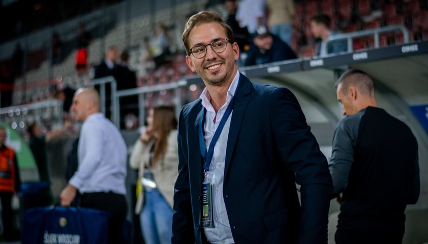 David Balda, dyrektor sportowy Śląska Wrocław, stoi uśmiechnięty przy ławce rezerwowych, z rękami w kieszeni, na nadgarstku ma zegarek.