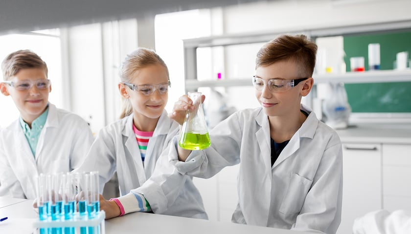 Dziewczynka i dwóch chłopców w białych fartuchach i okularach ochronnych podczas ćwiczeń laboratoryjnych, zdjęcie ilustracyjne 
