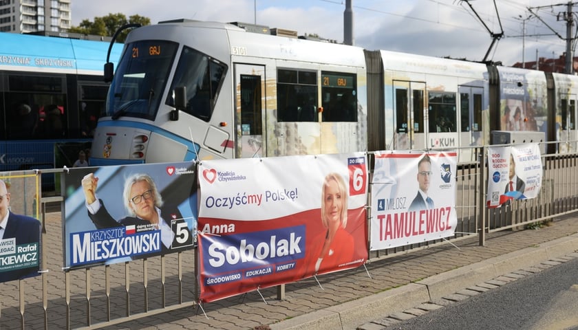 Plakaty wyborcze wiszące na płocie wzdłuż przystanku tramwajowego 