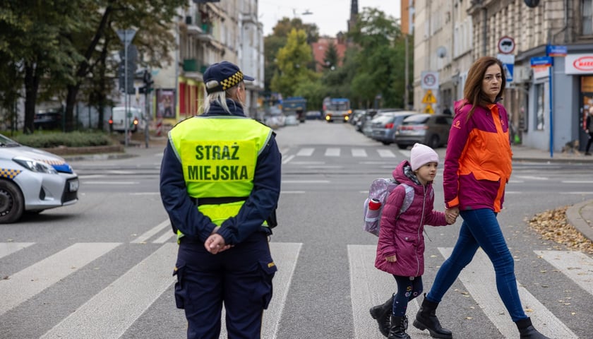 Na zdjęciu strażniczka miejska stoi przy przejściu dla pieszych, przez które przechodzi mama z córką za rękę