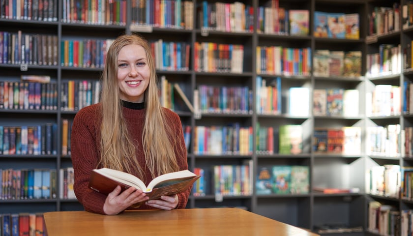 Uśmiechnięta kobieta z otwartą książką. W tle regały z książkami. Zdjęcie ilustracyjne wykonane w filii nr 12 Miejskiej Biblioteki Publicznej  przy ul. Piłsudskiego 105