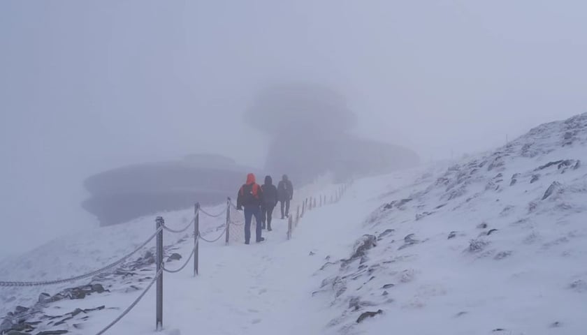  Trzy osoby idą pod górę. Szlak jest ośnieżony, na drugim planie widać zarys budynku Wysokogórskiego Obserwatorium Meteorologicznego na Śnieżce 