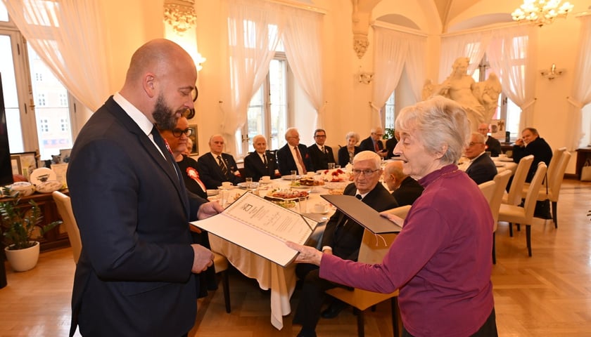 Prezydent Jacek Sutryk wręcza starszej kobiecie dyplom, w tle stół przy którym siedzą ludzie 
