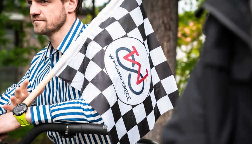 Mężczyzna w koszuli w biało-niebieskie paski trzyma flagę w czarno-białą szachownicę s logo W kółko kręcę