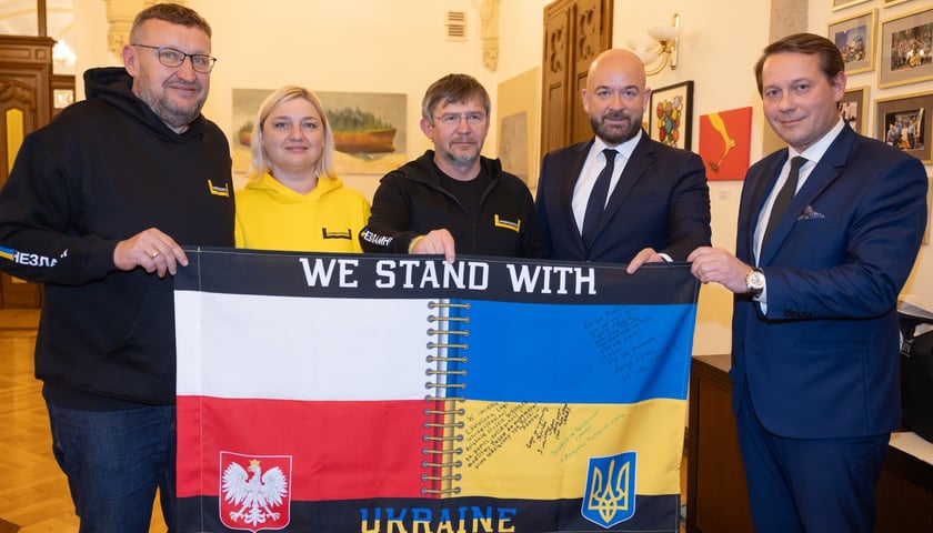 Prezydent Wrocławia Jacek Sutryk i delegacja ze Lwowa trzymają baner z flagami polską i ukraińską połączonymi szwem. Napis: We stand with Ukraine 
