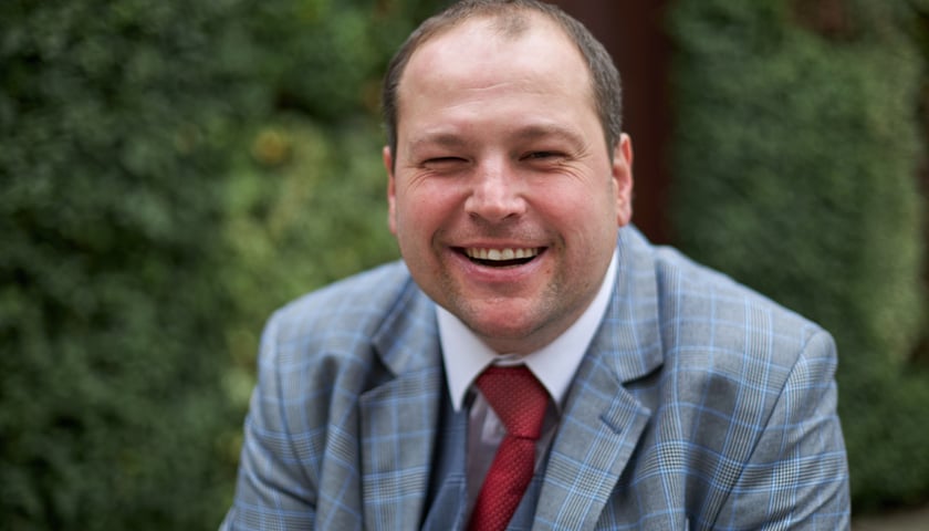 Uśmiechnięty mężczyzna w szarym garniturze - Michał Piechel społeczny doradca prezydenta Wrocławia do spraw osób z niepełnosprawnością
