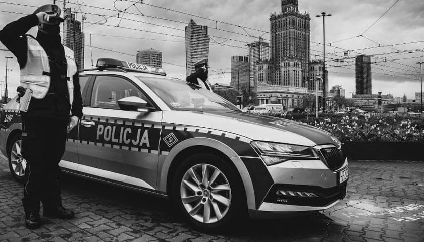 Czarnobiałe zdjęcie przedstawiające salutującego policjanta przy radiowozie.