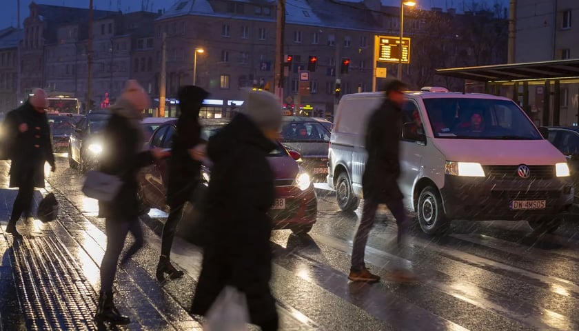Ludzie przechodzący przez ulicę po zmroku, pada deszcz 