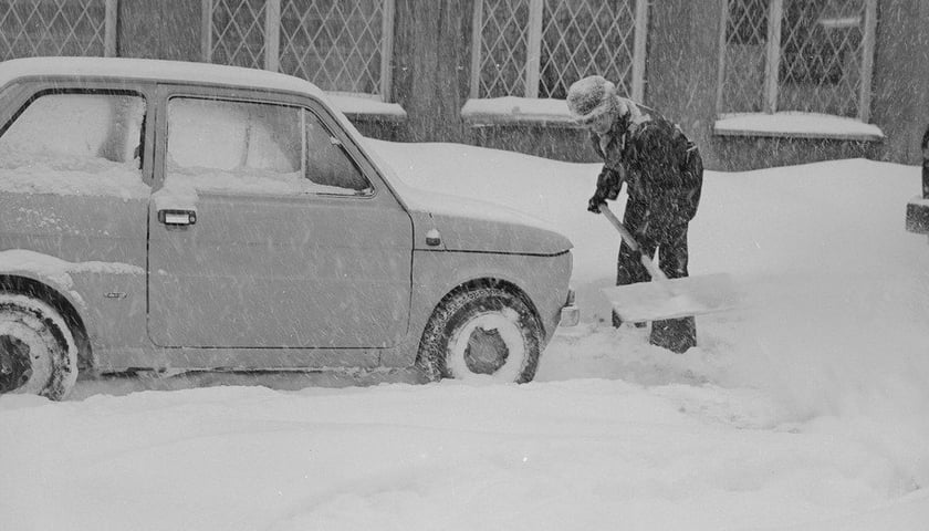 Mężczyzna z szuflą obok zasypanego śniegiem małego Fiata