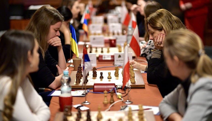 Rząd stołów z rozłożonymi szachownicami. Po obu stronach siedzą szachistki 