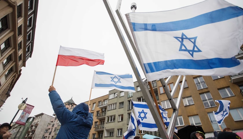 Mężczyzna wyznania mojżeszowego podnoszący nad głowę dwie flagi: Polski i Izraela. Widać także fragment tradycyjnej Chanukiji z zawieszoną flagą Izraela