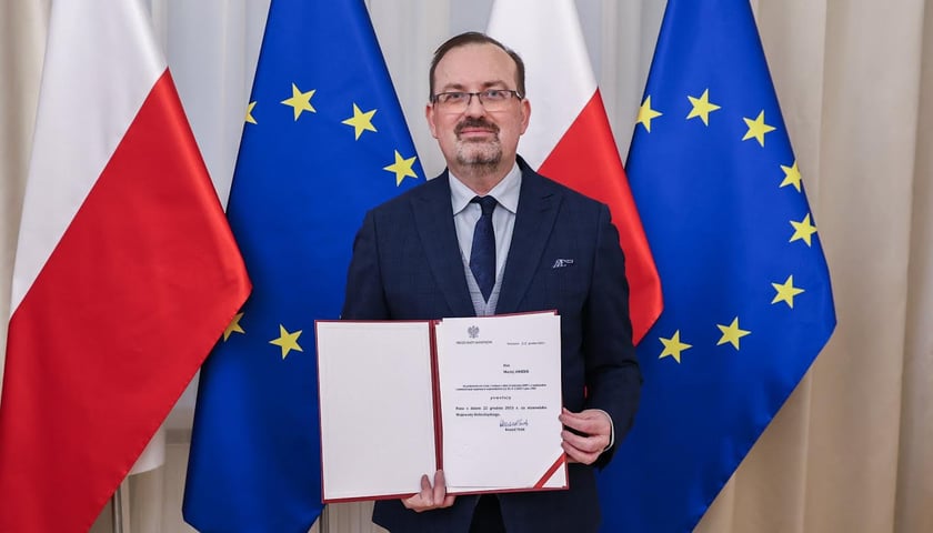 Mężczyzna w rękach trzyma dokument, w tle flagi Polski i Unii Europejskiej,  Maciej Awiżeń, wojewoda dolnośląski