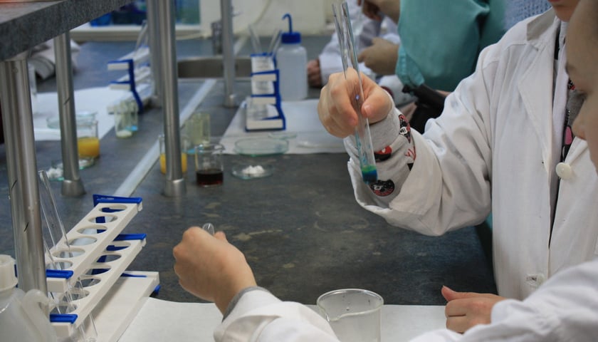 Stół w laboratorium, młode osoby ubrane w białe fartuchy trzymają w dłoni próbówki. Noc Biologów 2023