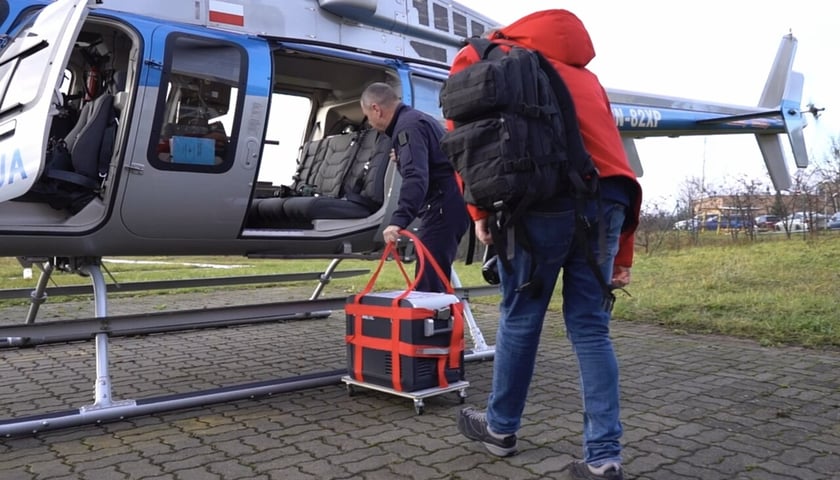 Policjant wkłada lodówkę z sercem przeznaczonym do transplantacji do policyjnego śmigłowca. Na pierwszym planie mężczyzna w czerwonej kurtce 