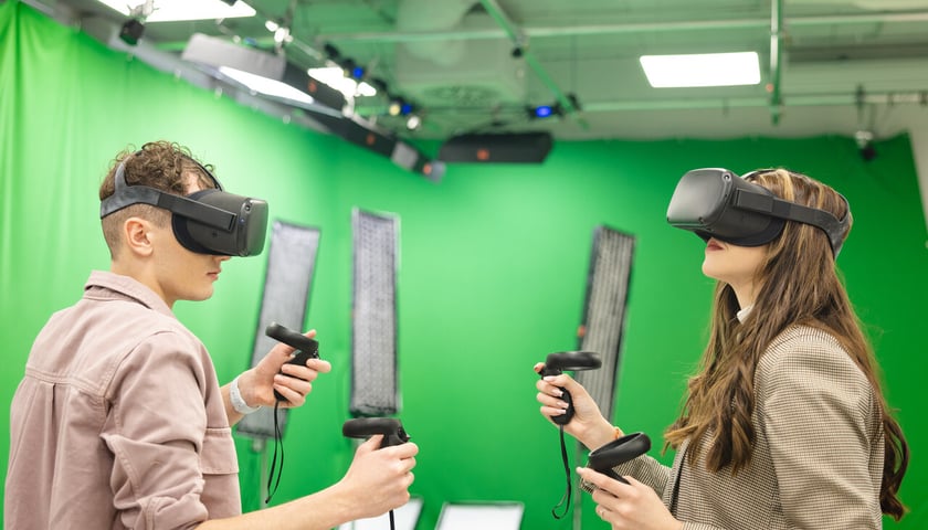 Dwie osoby z goglami do symulacji wirtualnej rzeczywistości stoją w pokoju o zielonych ścianach 