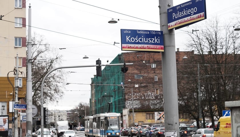 Słup z oznaczeniem ulic: Kościuszki i Pułaskiego, na dalszym planie: ulica Pułaskiego, tramwaj, auta