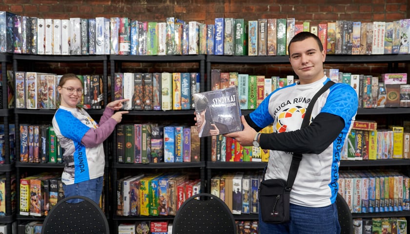 Dwoje przedstawicieli Stowarzyszenia Druga Runda przed regałem z grami planszowymi. Zdjęcie wykonane podczas festiwalu Wrocław Games Fest w Czasoprzestrzeni. Wrocław, listopad 2023.