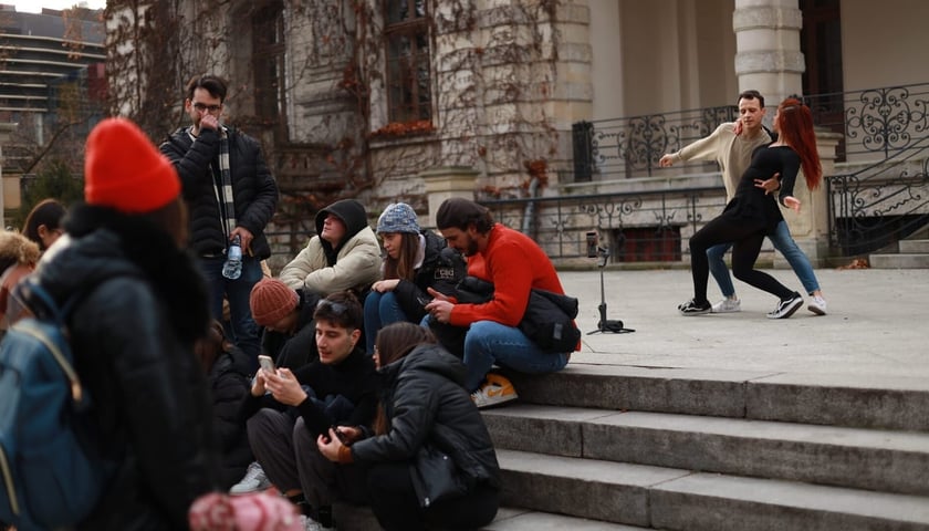 Z lewej strony grupka młodych ludzi siedzących na schodkach w miejskim parku. Z prawej strony, na drugim planie, para tancerzy. Zdjęcie ilustracyjne.