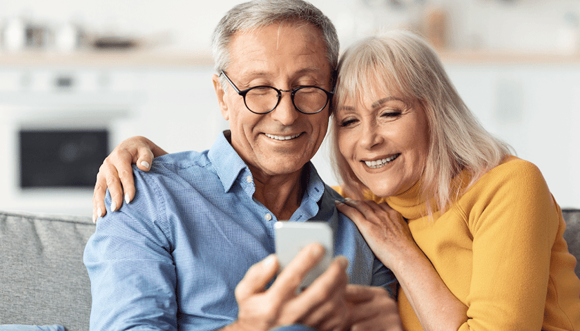 Starszy mężczyzna w okularach i obejmująca go kobieta patrzą na telefon komórkowy
