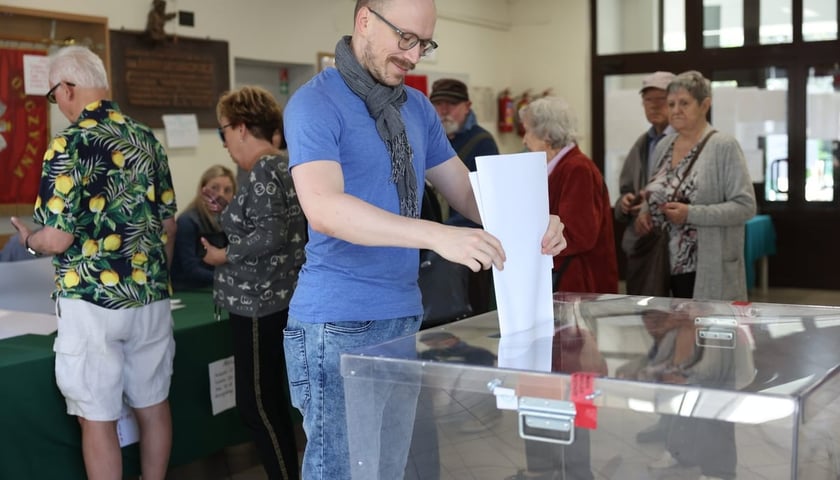 Mężczyzna w okularach wrzuca karty do głosowania do urny wyborczej, w tle widać innych głosujących