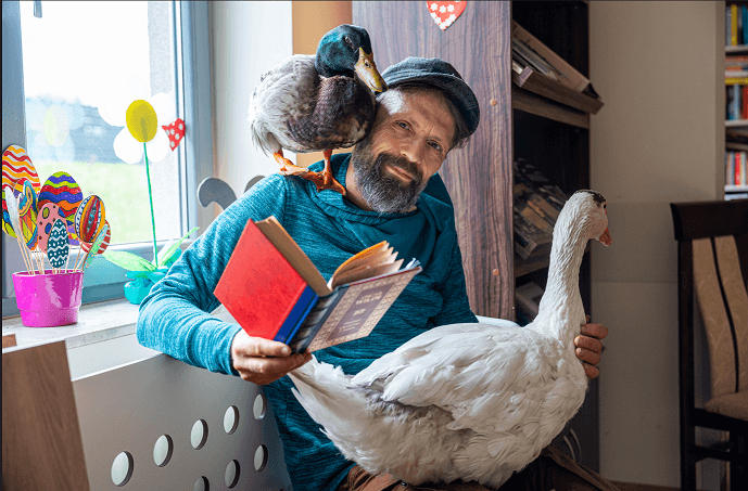 Dariusz Brzeski w czapce z daszkiem i niebieskiej bluzie z kaczorem Wirkiem na ramieniu, gęsią Sunny na kolanach i książką w ręce