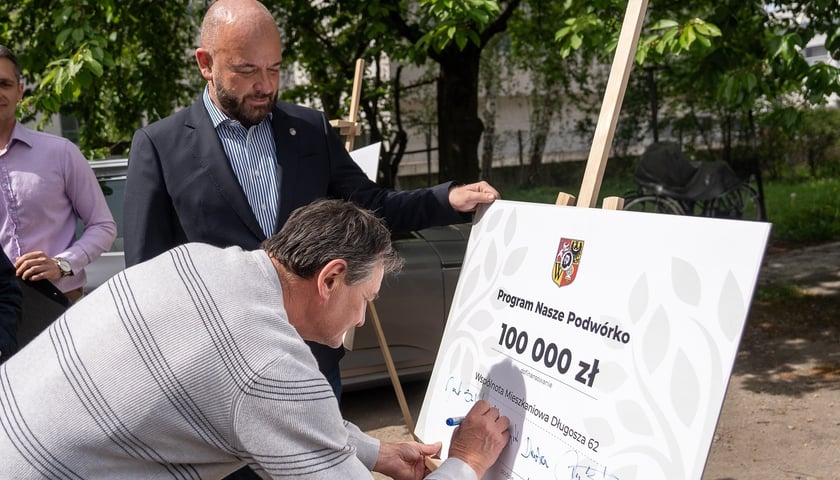 Mieszkaniec składa podpis na planszy z napisem: Program Nasze Podwórko 100 000 zł dofinansowania. Wspólnota Mieszkaniowa Długosza 62. Obok stoi prezydent Wrocławia Jacek Sutryk 