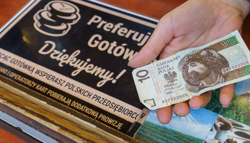 <p>Dłoń z banknotem 10-złotowym na tle informacji na stoisku w Hali Targowej: Preferujemy got&oacute;wkę. Dziękujemy! Płacąc got&oacute;wką wspierasz polskich przedsiębiorc&oacute;w. Banki i opretorzy kart pobierają dodatkową prowizję</p>